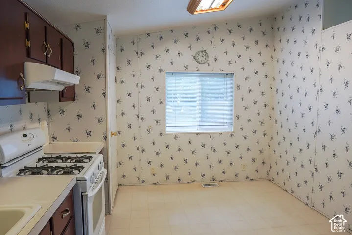 Brown wood-finish cabinetry, white countertop, white gas range, and a window with horizontal blinds