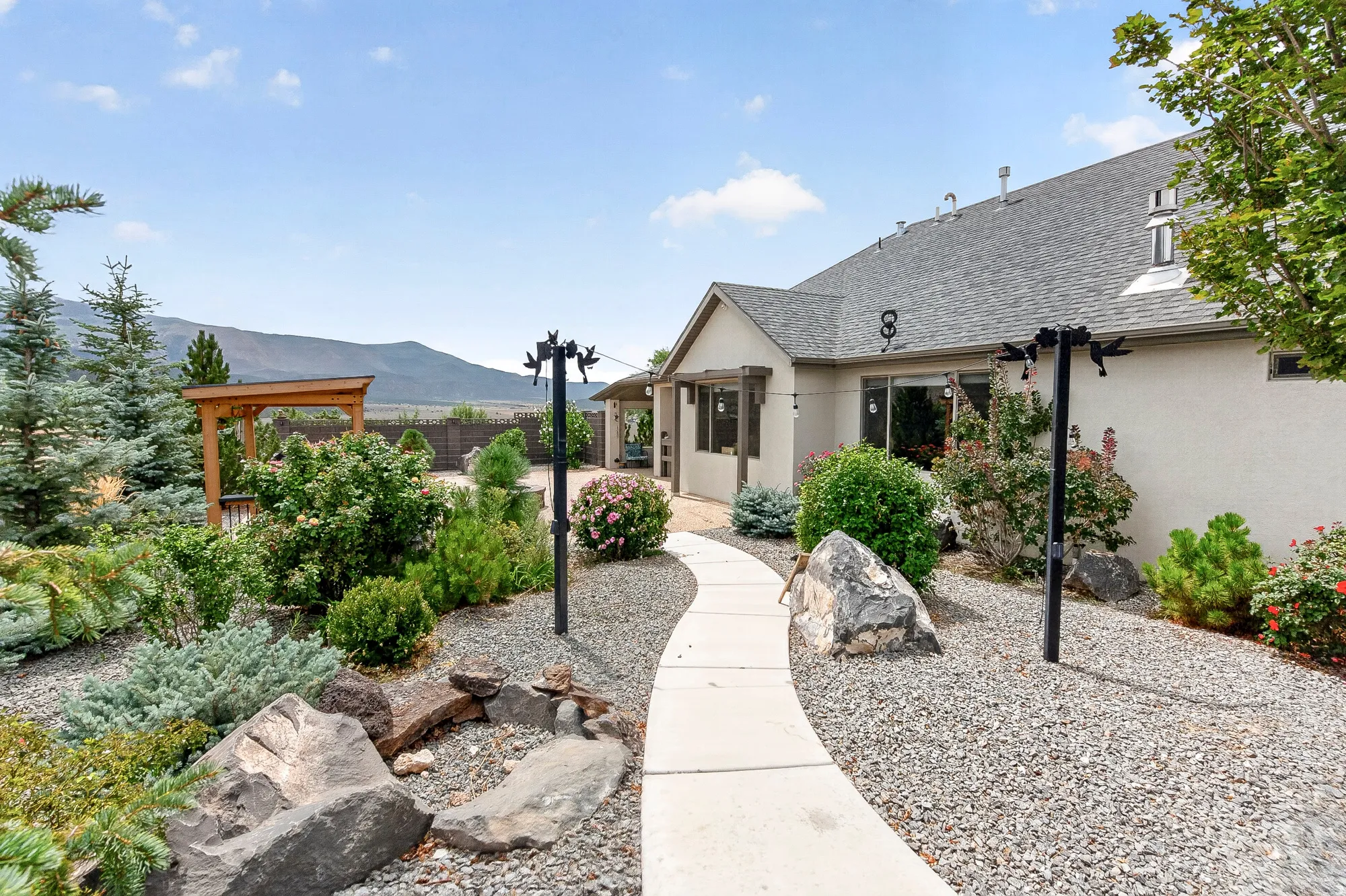 Welcoming entry courtyard featuring a curved concrete walkway, mature landscaping with native plantings and specimen boulders, wooden pergola structure, and mountain backdrop