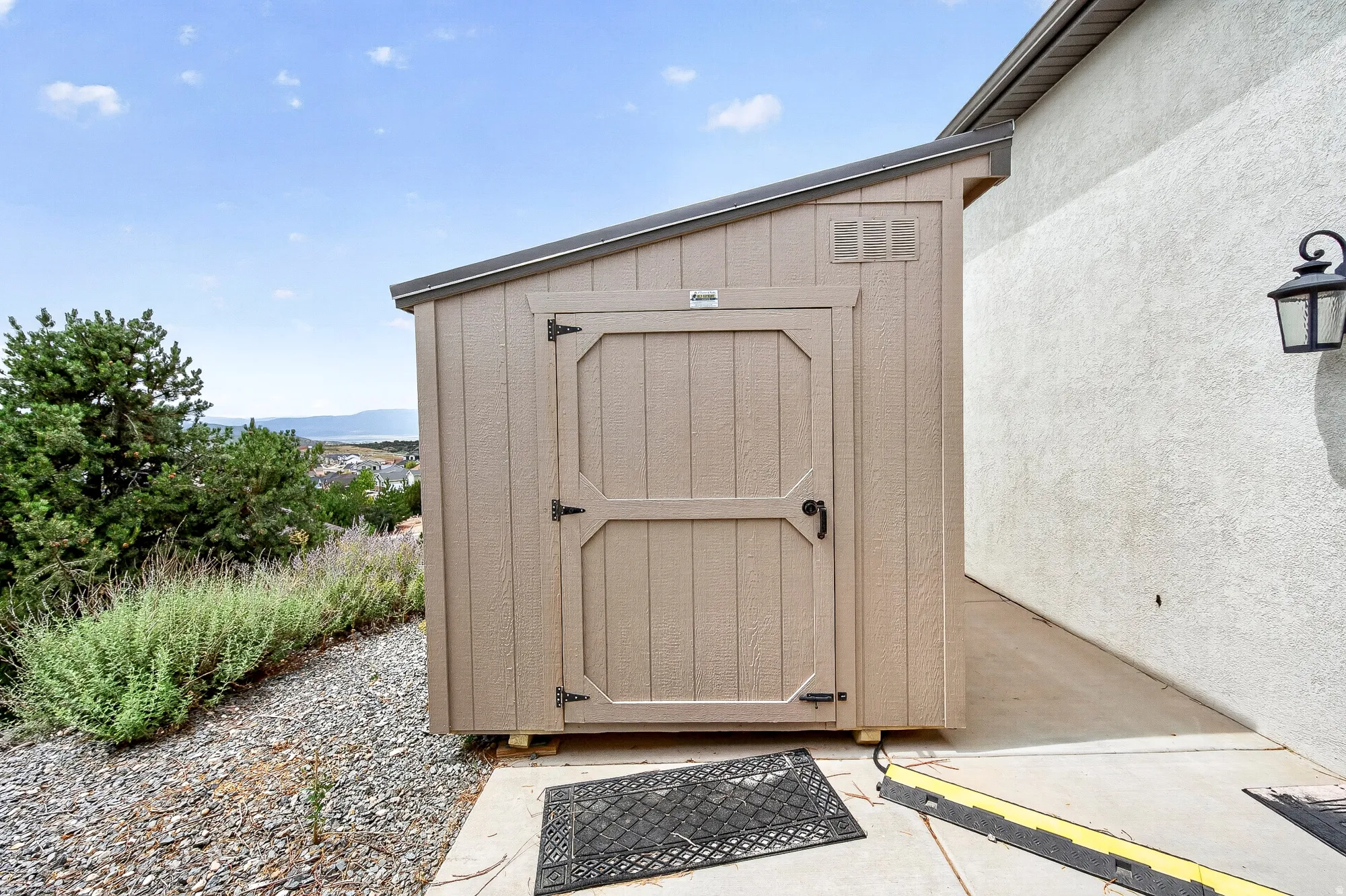 Exterior storage shed with a lean-to roof, situated on a concrete pad, adjacent to a stucco wall with an outdoor light fixture