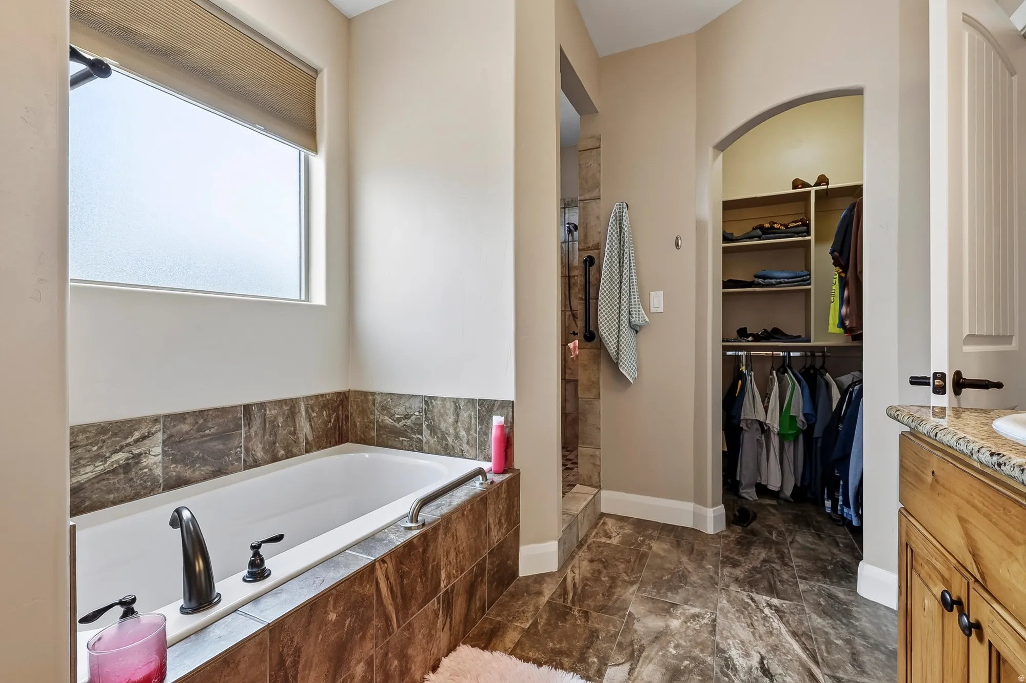 Spa-inspired primary bathroom featuring a soaking tub with tile surround, dual vanity with wood cabinetry and stone countertops, walk-in closet with built-in shelving and clothing rods, and large window providing natural light