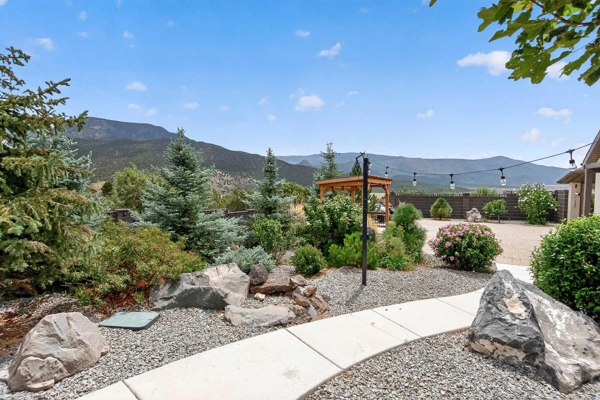 Professionally landscaped outdoor living space featuring a concrete pathway, river rock hardscaping, mature evergreen plantings, and a wooden pergola structure