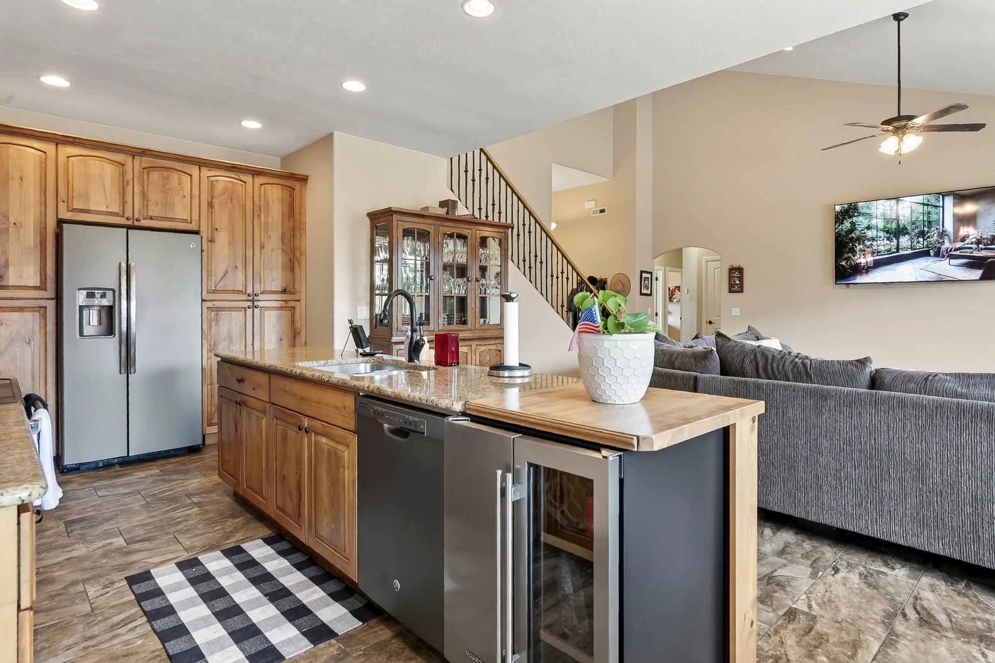 Open-concept kitchen and living area featuring custom wood cabinetry, stainless steel appliances including a side-by-side refrigerator, large center island with black base and wood countertop, wood-finish flooring, and recessed lighting throughout