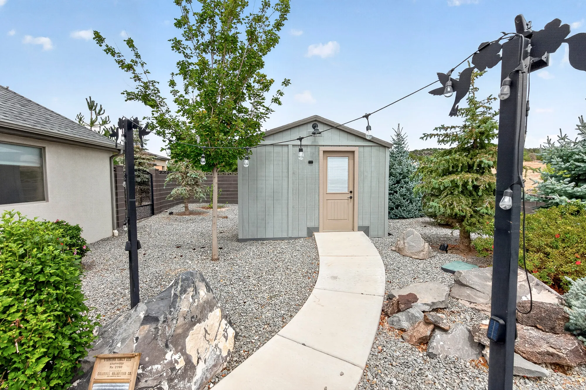 Modern backyard retreat featuring a contemporary garden shed with neutral exterior finishes, concrete pathway, gravel landscaping, mature tree plantings, and integrated landscape lighting throughout the grounds