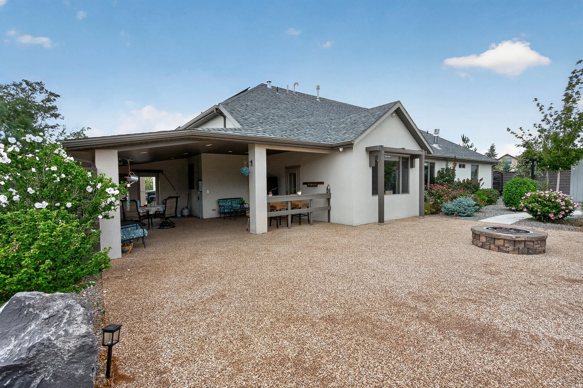 Expansive gravel courtyard with mature landscaping and established shrubs, anchoring a single-story residence featuring a covered patio with white columns, composite roofing, and multiple glass doors opening to the outdoor space