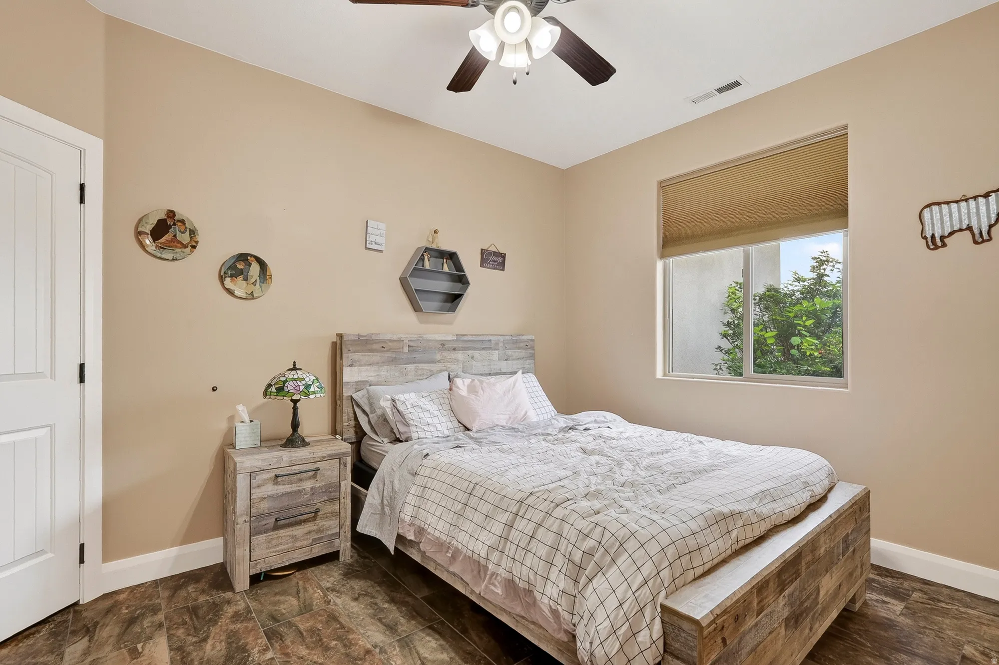 Spacious bedroom featuring a wood-finish bed frame with storage drawer base, coordinating wood-finish nightstand, ceiling fan with integrated lighting, and large picture window with cellular shade