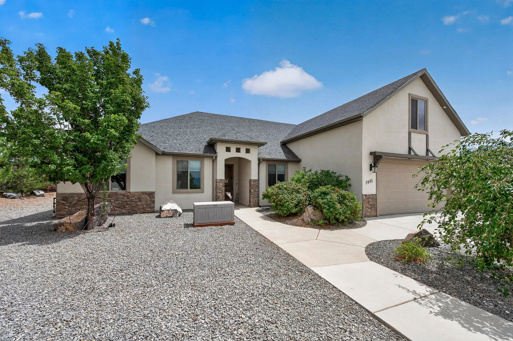 Contemporary home featuring a stucco and brick exterior with a gabled roofline, complemented by mature landscaping and xeriscaped grounds with decorative stone hardscaping