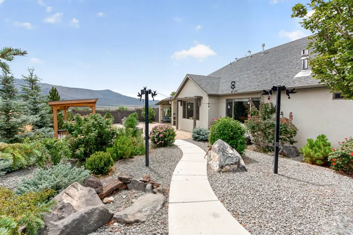 Welcoming entry courtyard featuring a curved concrete walkway, mature landscaping with native plantings and specimen boulders, wooden pergola structure, and mountain backdrop