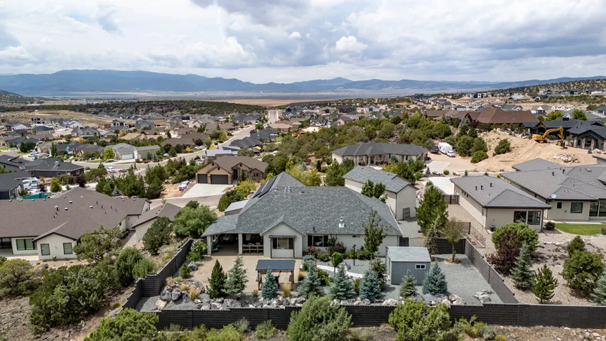 Aerial view showcasing a modern residence with a distinctive peaked roof and dark asphalt shingles, surrounded by professionally landscaped grounds featuring mature trees and manicured lawn
