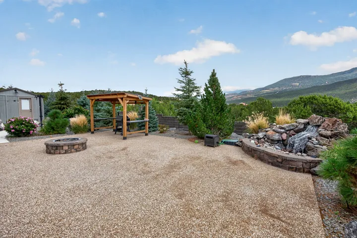 Gravel patio area featuring a stone fire pit, a wooden pergola, and a rock waterfall feature with a stone retaining wall