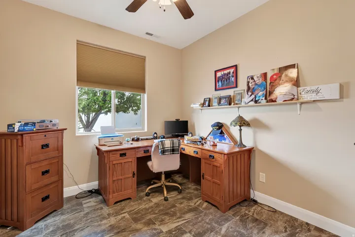 Dedicated home office featuring an L-shaped wood-finish desk with multiple storage drawers, a cushioned task chair, and abundant natural light from a large window with cellular shade