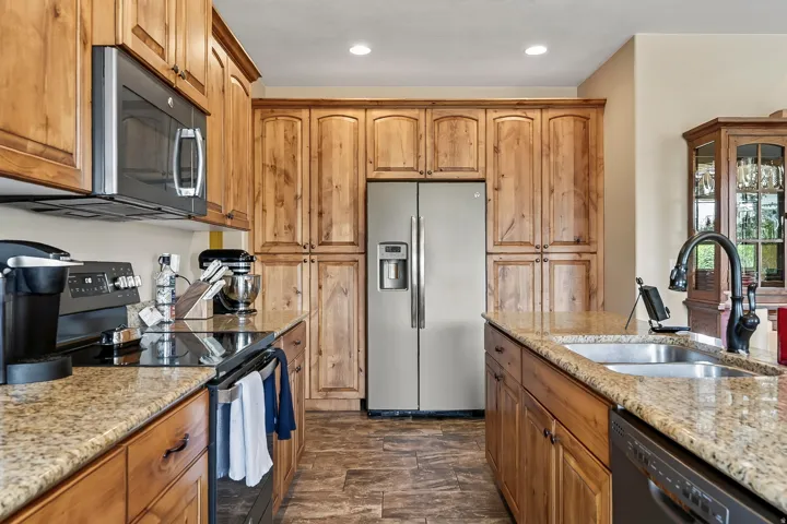 Galley-style kitchen featuring warm wood cabinetry with arched door fronts, stainless steel side-by-side refrigerator with ice and water dispensers, granite countertops, and dual undermount sinks with bronze faucetry