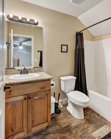 Spa-inspired bathroom featuring a wood-finish vanity with dual-handle faucet and granite-topped countertop, framed mirror with integrated lighting, white ceramic toilet, and tile surround shower with dark curtain rod