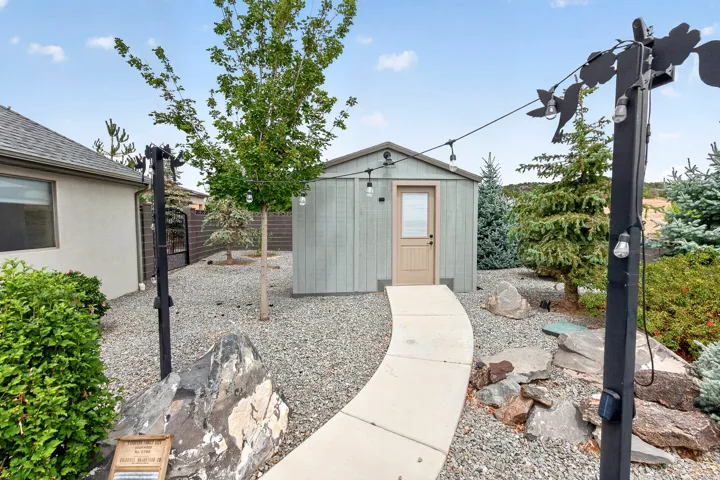 Modern backyard retreat featuring a contemporary garden shed with neutral exterior finishes, concrete pathway, gravel landscaping, mature tree plantings, and integrated landscape lighting throughout the grounds