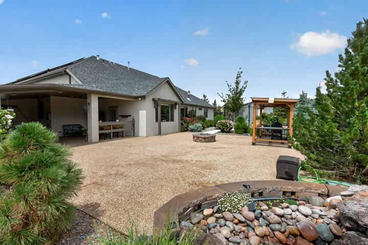 Spacious backyard retreat featuring a generous gravel entertaining area, mature landscaping with native plantings, decorative fire pit with stone surround, wooden pergola structure, and a single-story residence with covered patio and gabled roofline