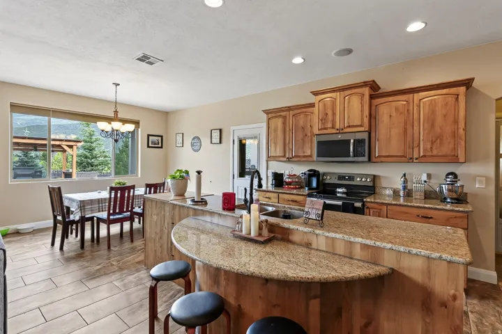 Open-concept kitchen and dining area featuring custom wood cabinetry, granite-topped curved island with seating, stainless steel appliances, tile flooring, and abundant natural light from expansive windows overlooking the landscape