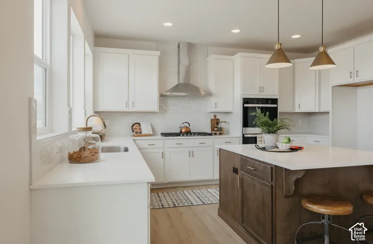 White shaker cabinetry, light wood-finish flooring, stainless steel range hood, dual wall ovens, and a wood-finish island with seating