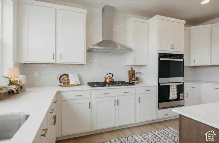 Modern kitchen featuring white shaker cabinetry, stainless steel range hood, built-in double oven, and wood-finish flooring