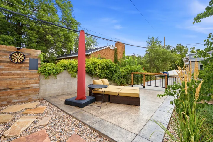 Stamped concrete patio with a horizontal wood-paneled accent wall, a retaining wall featuring lush greenery, and a black wrought iron fence with a wood top rail