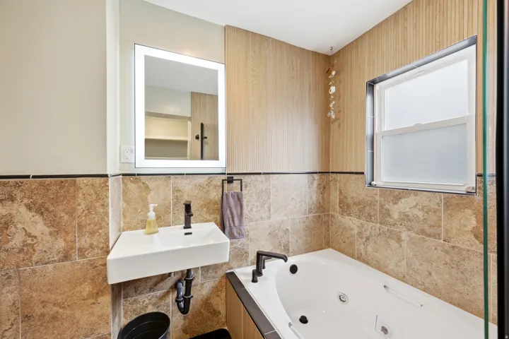 Soaking tub with dark fixtures, wall-mounted sink with dark faucet, back-lit mirror, and tile wainscoting