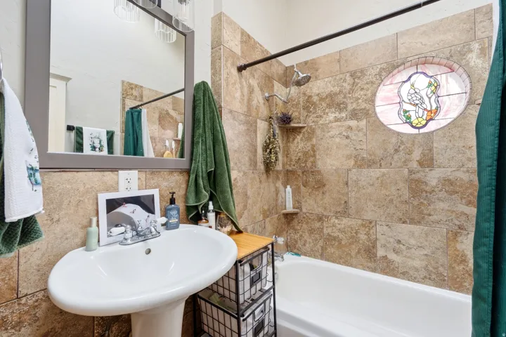 Pedestal sink with chrome faucet, large framed mirror, tiled shower surround, built-in shower shelf, and circular stained glass window