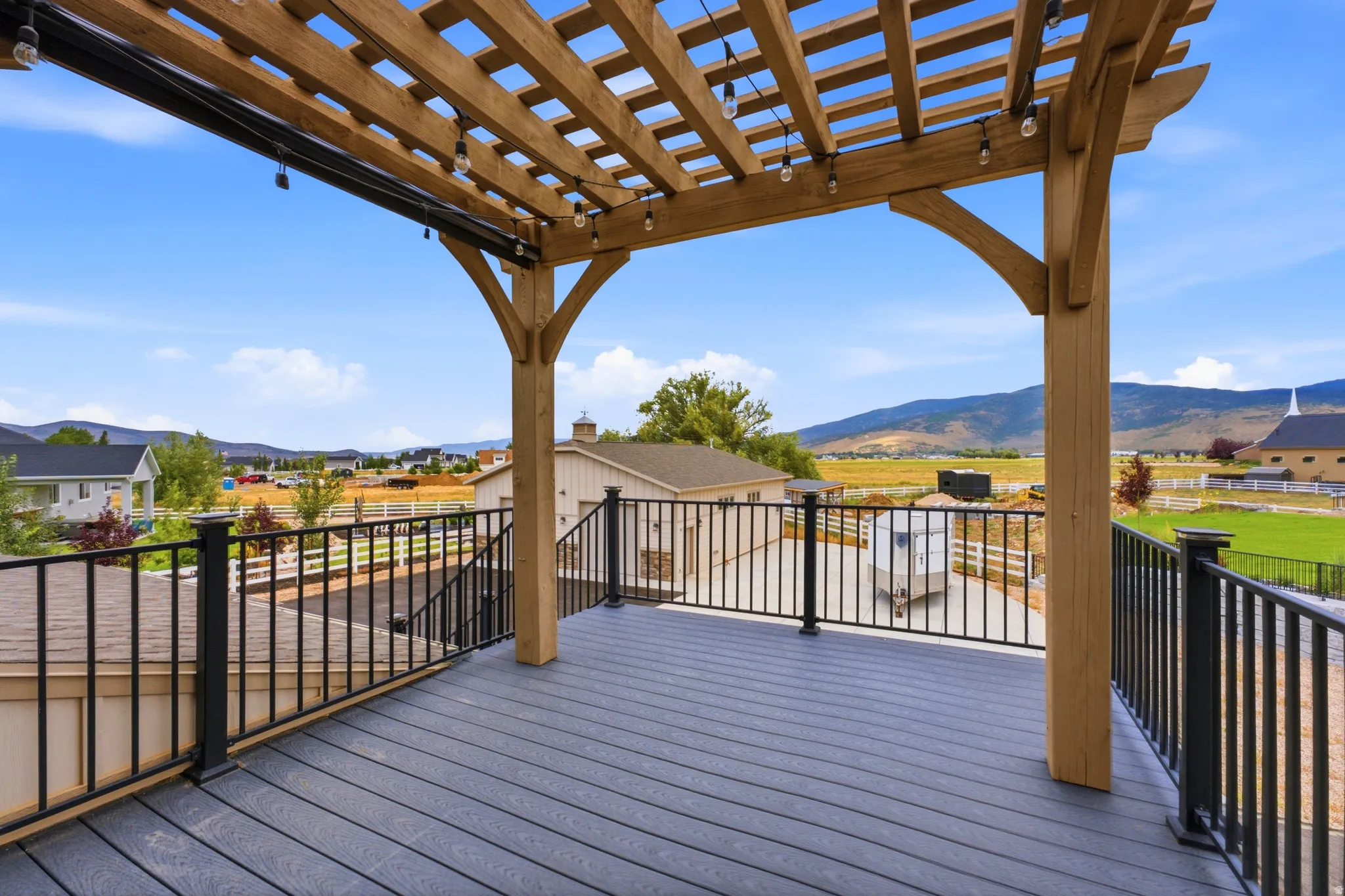 Expansive composite deck with wood-finish flooring and overhead pergola structure, featuring black metal railings and sweeping vistas of rolling hills and mountain landscape beyond neighboring properties
