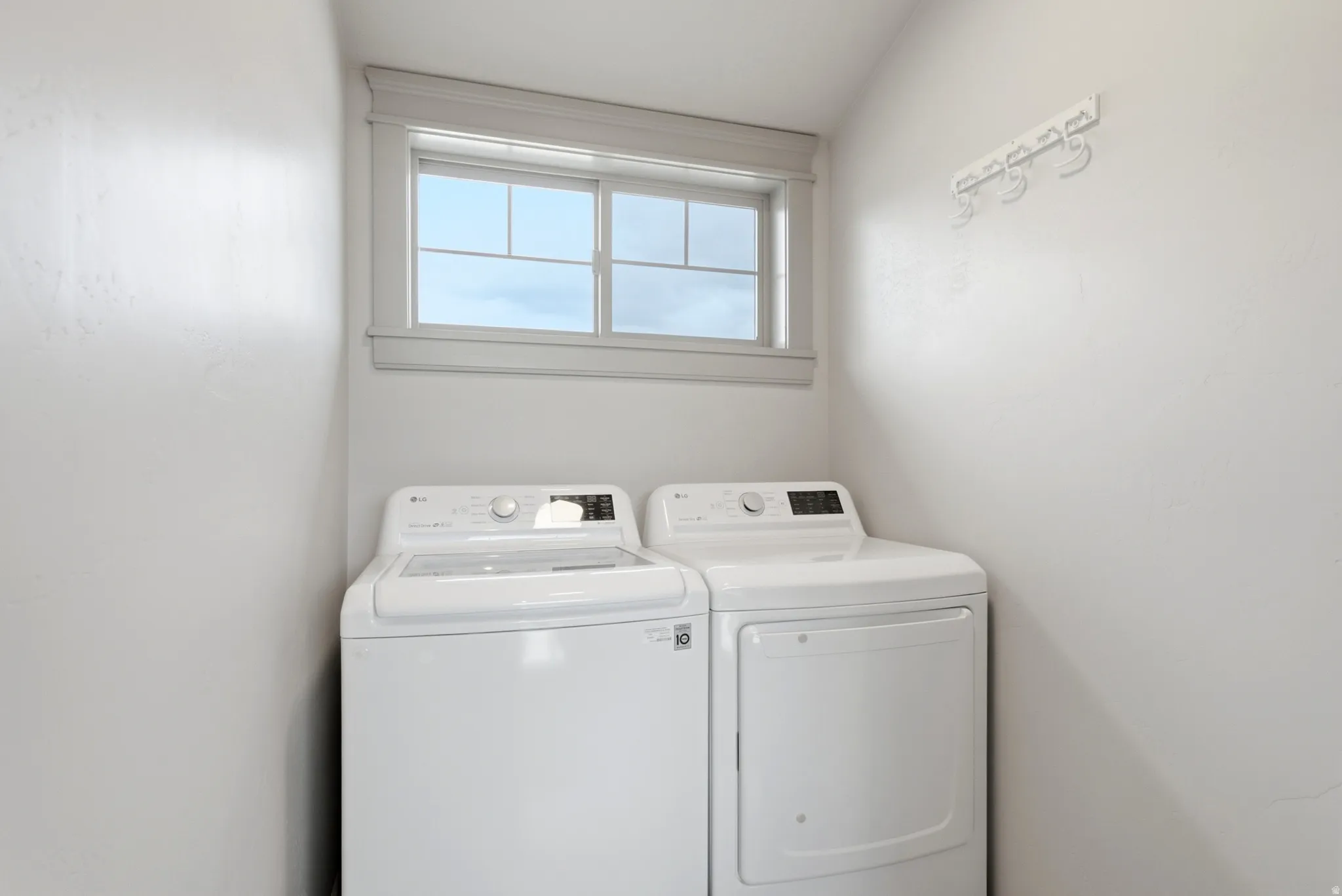 Dedicated laundry room featuring side-by-side washer and dryer with modern control panels, bright natural light from a fixed window, and clean white finishes throughout the compact space