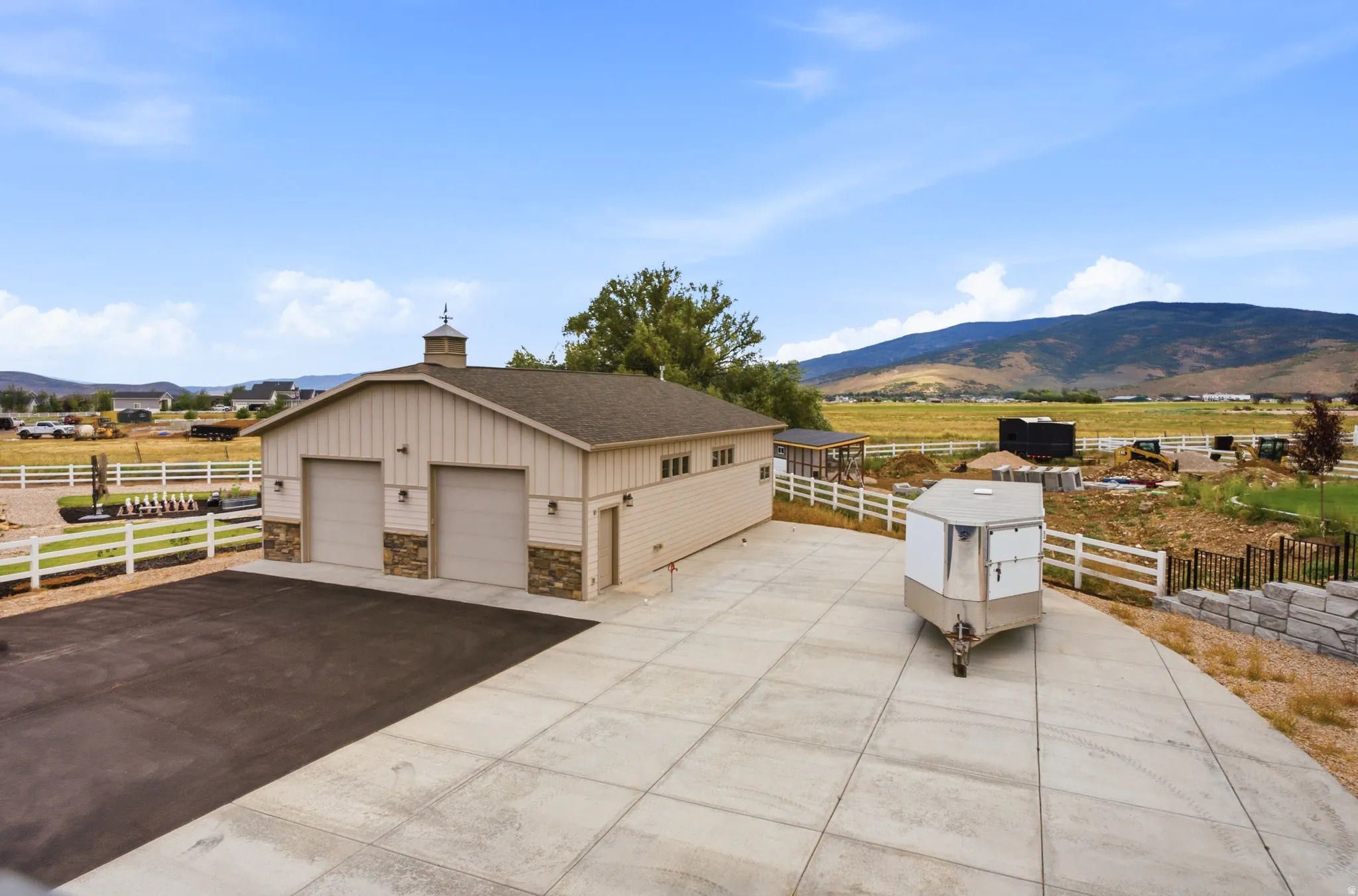 Spacious agricultural facility featuring a tan metal-frame structure with multiple garage bays, concrete apron, white rail fencing, and an attached horse trailer