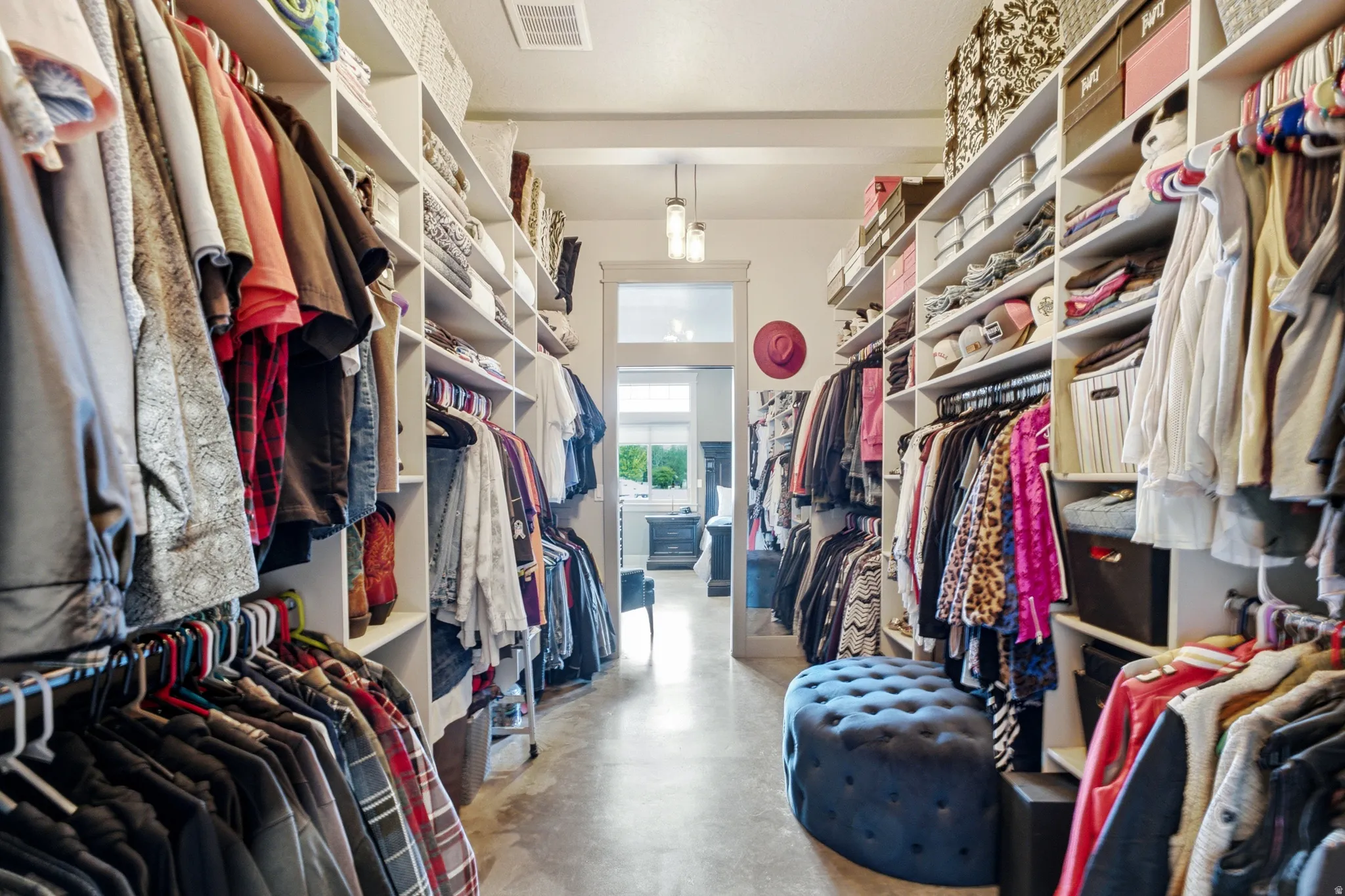 Spacious walk-in closet featuring extensive built-in shelving and clothing rods throughout, polished concrete flooring, recessed lighting, and a blue upholstered ottoman