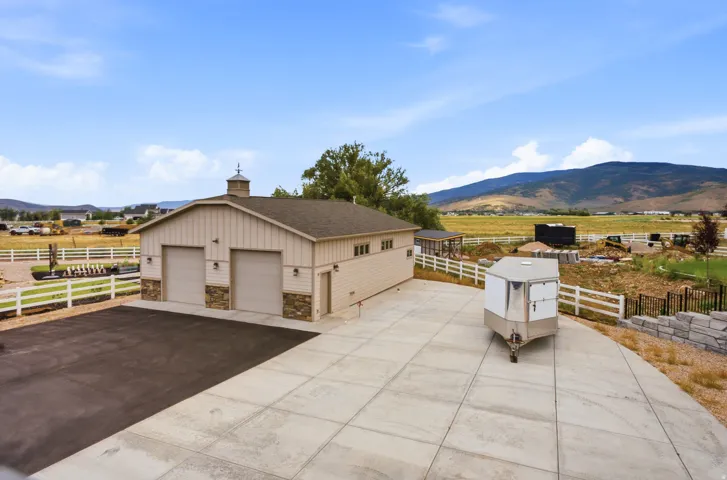 Spacious agricultural facility featuring a tan metal-frame structure with multiple garage bays, concrete apron, white rail fencing, and an attached horse trailer