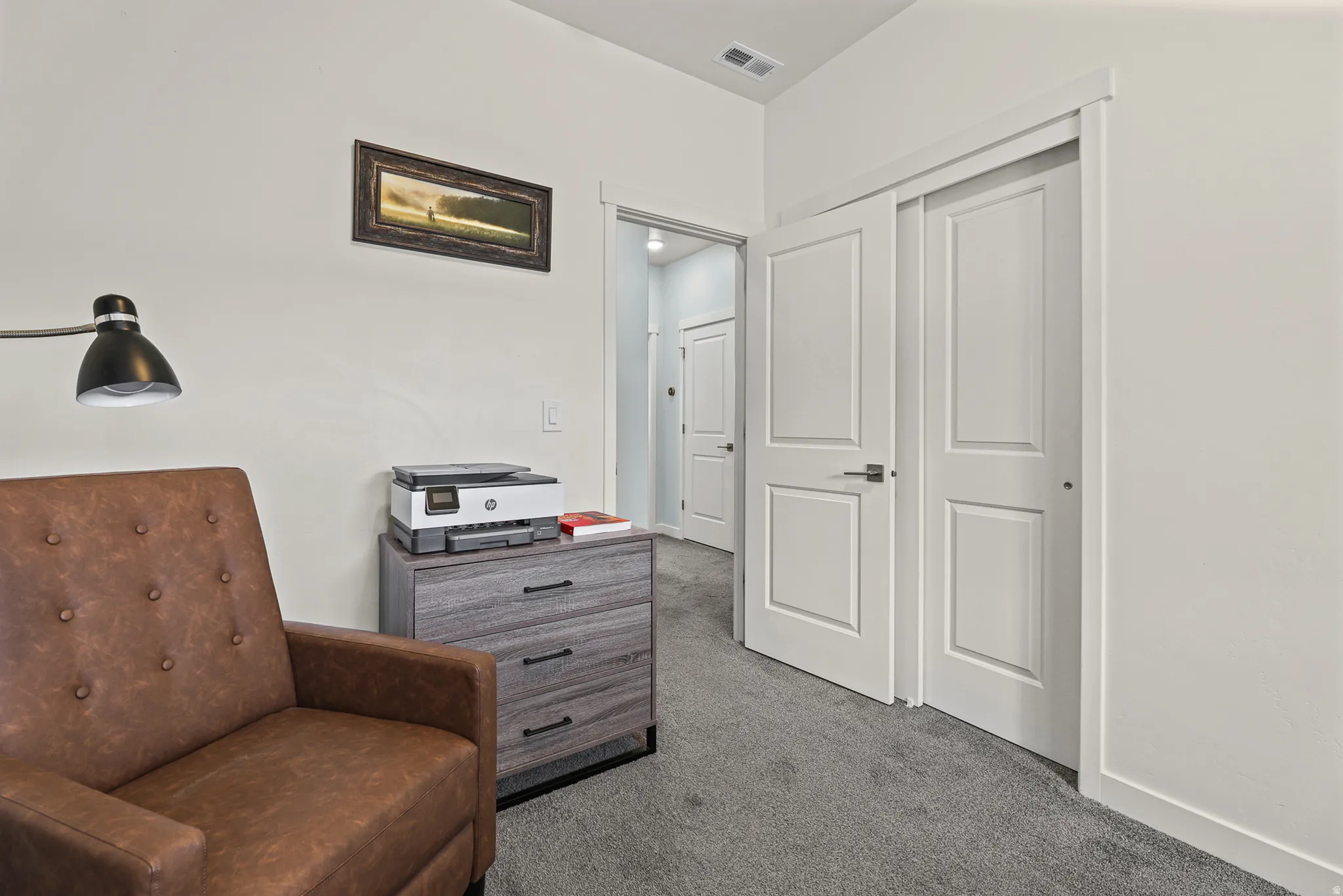 Bedroom or office space featuring carpet flooring, white six-panel closet doors, gray wood-finish dresser with storage, black wall-mounted task lighting, and white walls with crown molding
