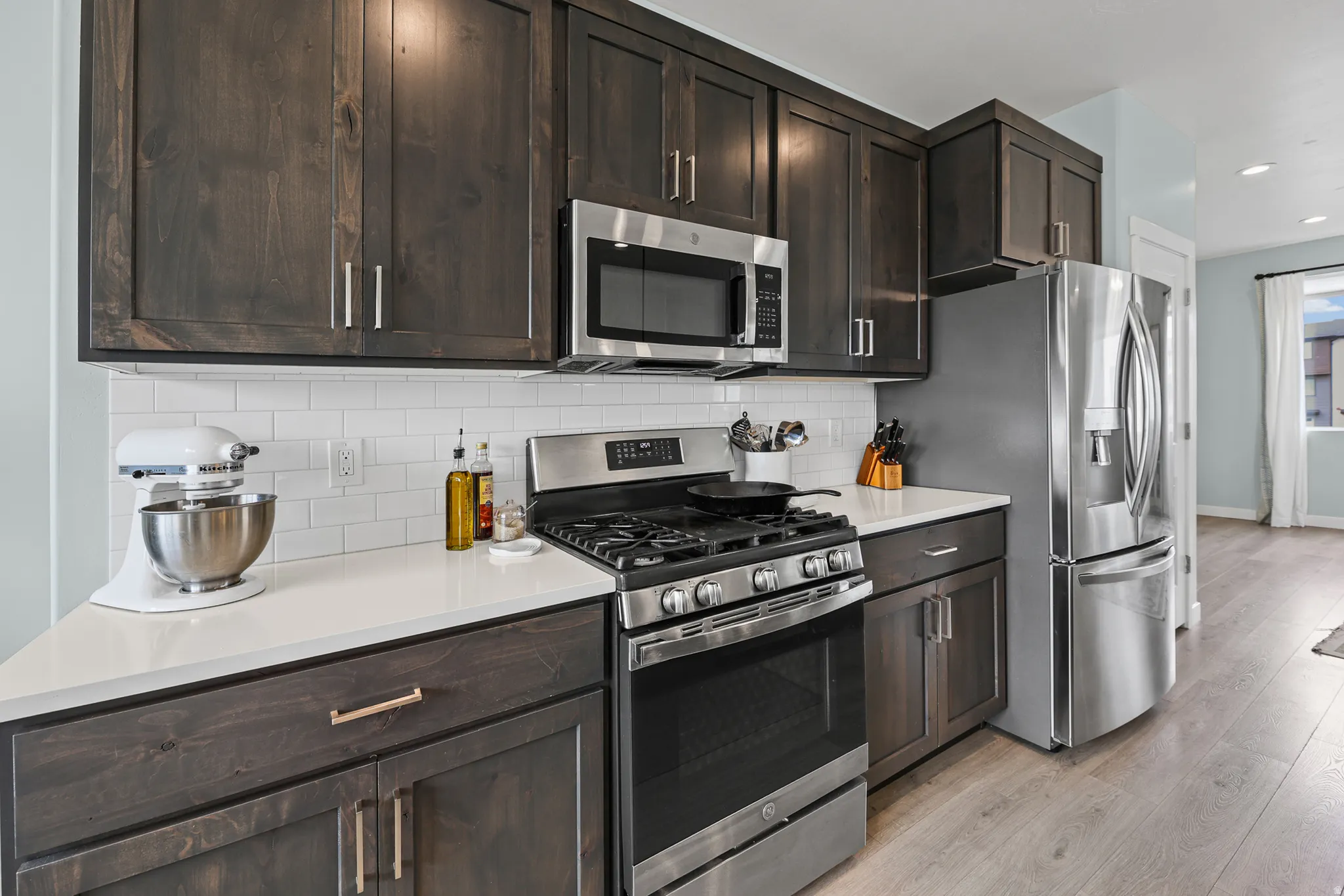 Dark wood-finish cabinetry, white subway tile backsplash, light-toned countertops, stainless steel appliances, and wood-finish flooring