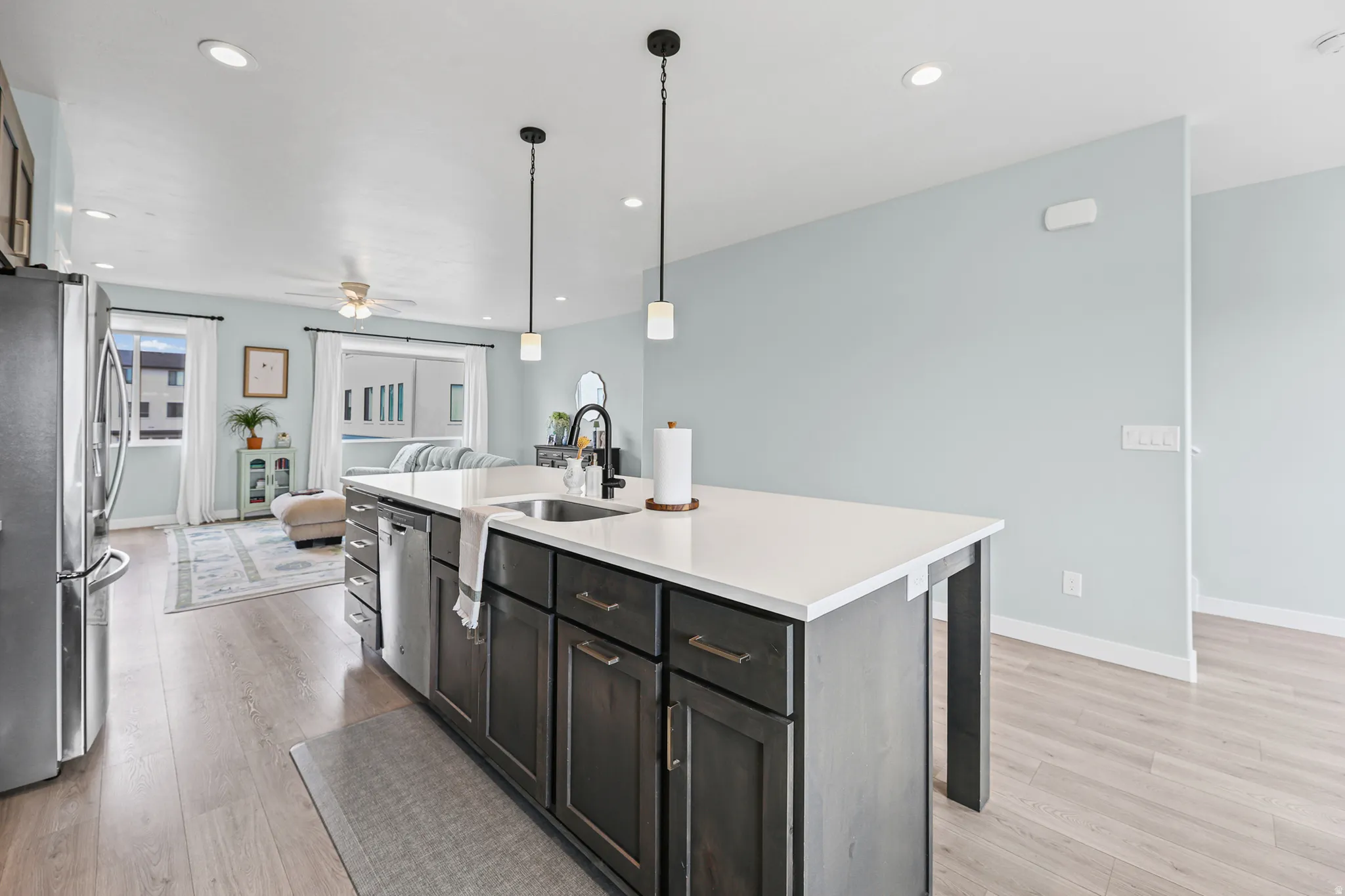 Modern kitchen featuring a spacious island with white quartz countertop and dark cabinetry, stainless steel appliances including a professional-grade refrigerator, pendant lighting with matte black fixtures, and wood-finish flooring throughout the open-co