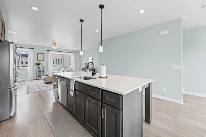 Modern kitchen featuring a spacious island with white quartz countertop and dark cabinetry, stainless steel appliances including a professional-grade refrigerator, pendant lighting with matte black fixtures, and wood-finish flooring throughout the open-co