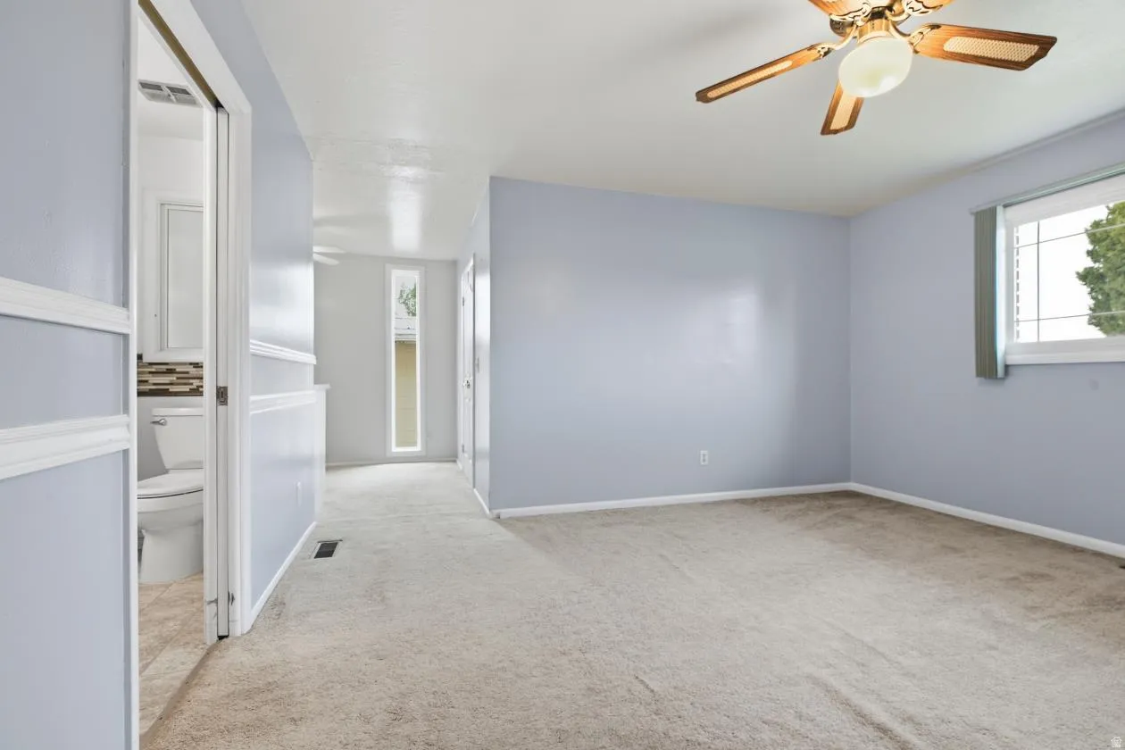Carpeted flooring, ceiling fan with integrated lighting, natural light from a window, and an adjacent bathroom with tiled flooring and a tiled backsplash