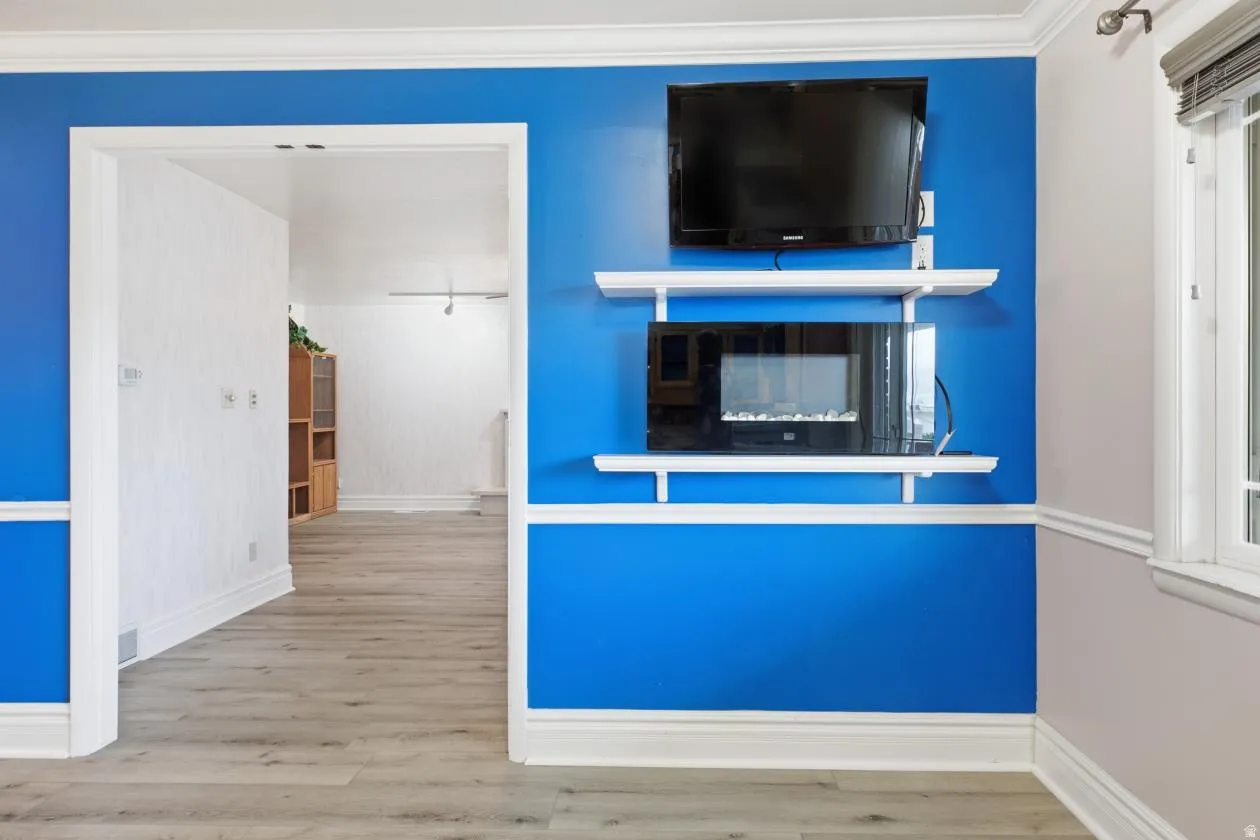 Wood-finish flooring, accent wall with chair rail molding, built-in electric fireplace, white crown molding, and window with blinds