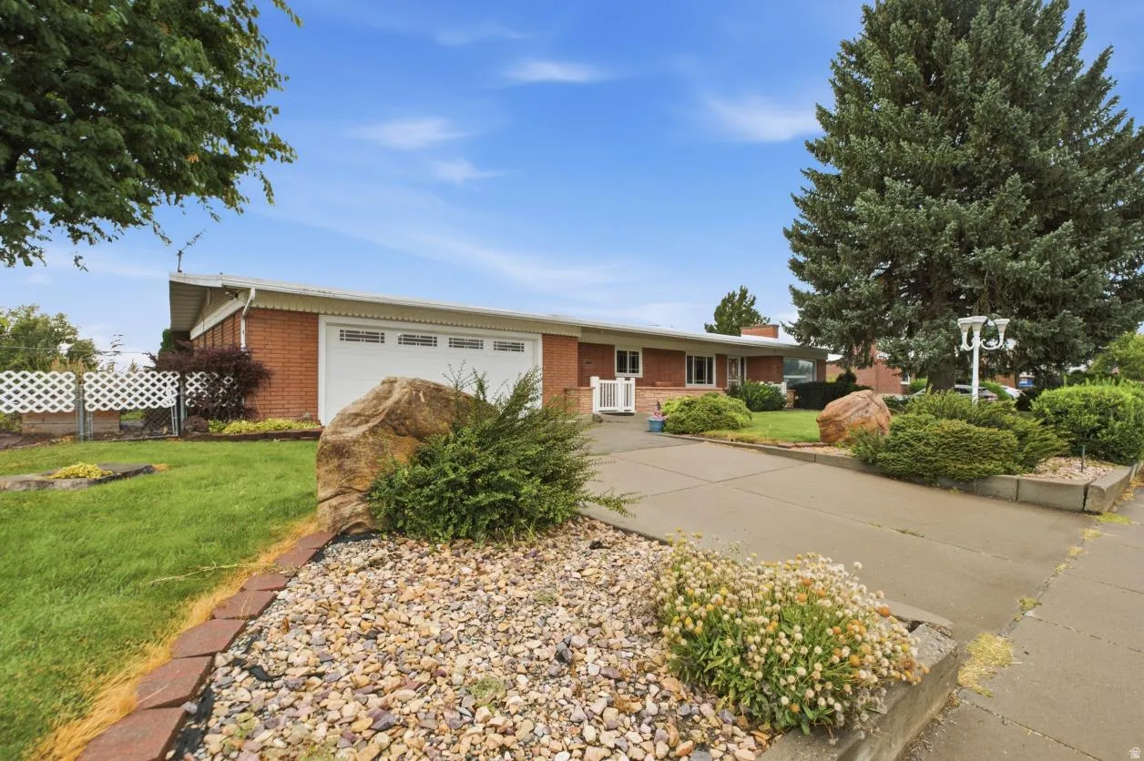 Brick exterior featuring a two-car garage with white door, concrete driveway, and mature landscaping with decorative rock beds