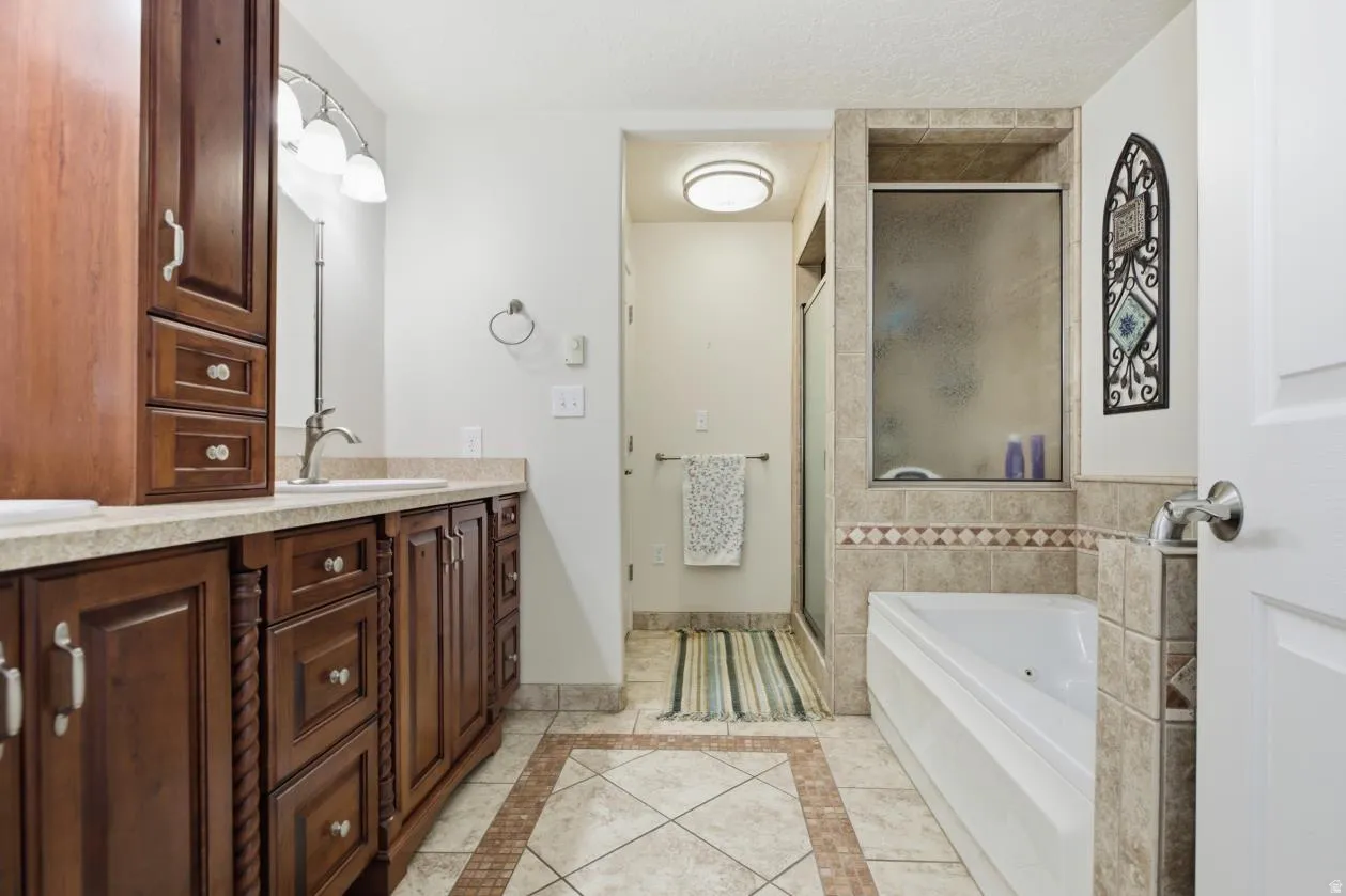 Dual sink vanity with dark wood cabinetry and light countertops, a built-in shower, soaking tub, and tile flooring