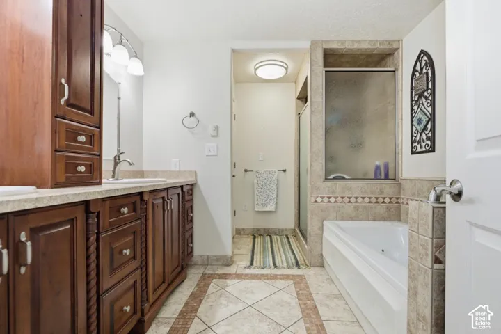 Dual sink vanity with dark wood cabinetry and light countertops, a built-in shower, soaking tub, and tile flooring