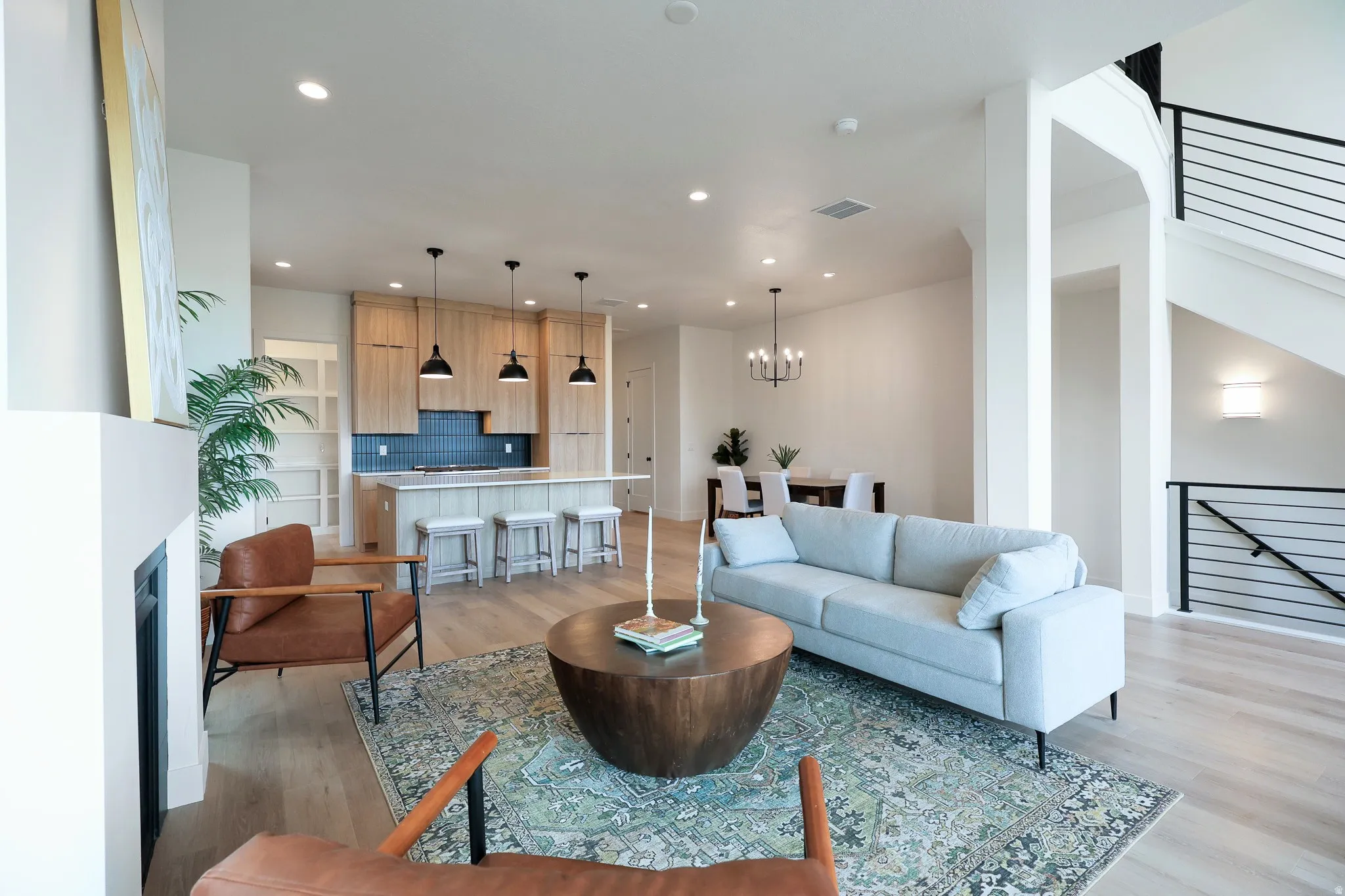 Open-concept living area featuring light wood-finish flooring, a modern kitchen with a blue subway tile backsplash, a kitchen island with pendant lighting, a built-in fireplace, and a staircase with contemporary black railings