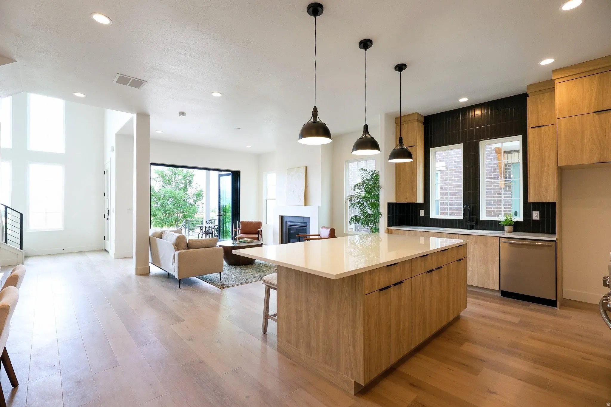 Open-concept living area featuring light wood-finish flooring, a kitchen island with a light-toned countertop, black tile backsplash, and stainless steel appliances