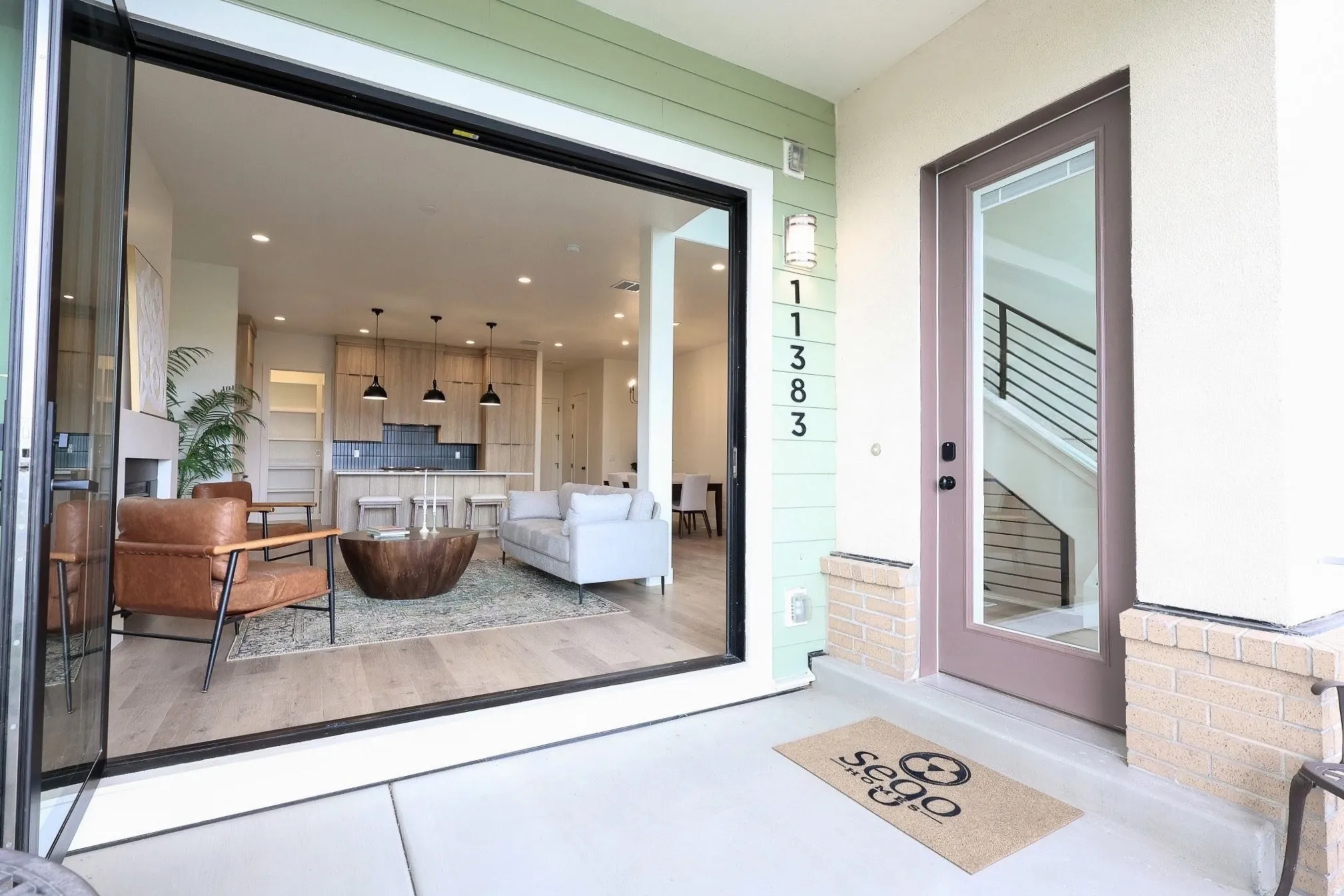 Expansive glass bi-fold doors open to an interior with wood-finish flooring, a contemporary kitchen with a dark tile backsplash, and light wood cabinetry