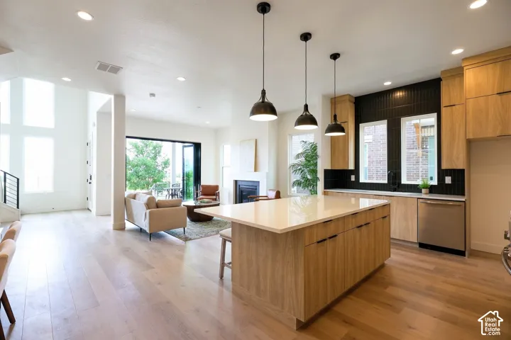 Open-concept living area featuring light wood-finish flooring, a kitchen island with a light-toned countertop, black tile backsplash, and stainless steel appliances
