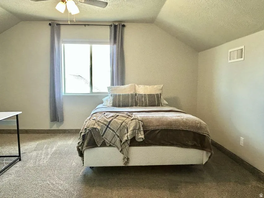 Sloped ceiling, window with curtain rod, ceiling fan with light fixture, carpeted flooring, and a wall-mounted vent