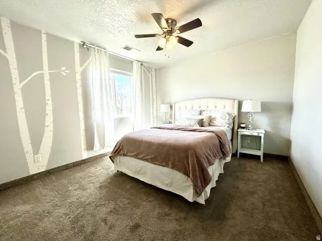 Carpeted bedroom with a ceiling fan light fixture, a window with sheer drapes, and an upholstered headboard