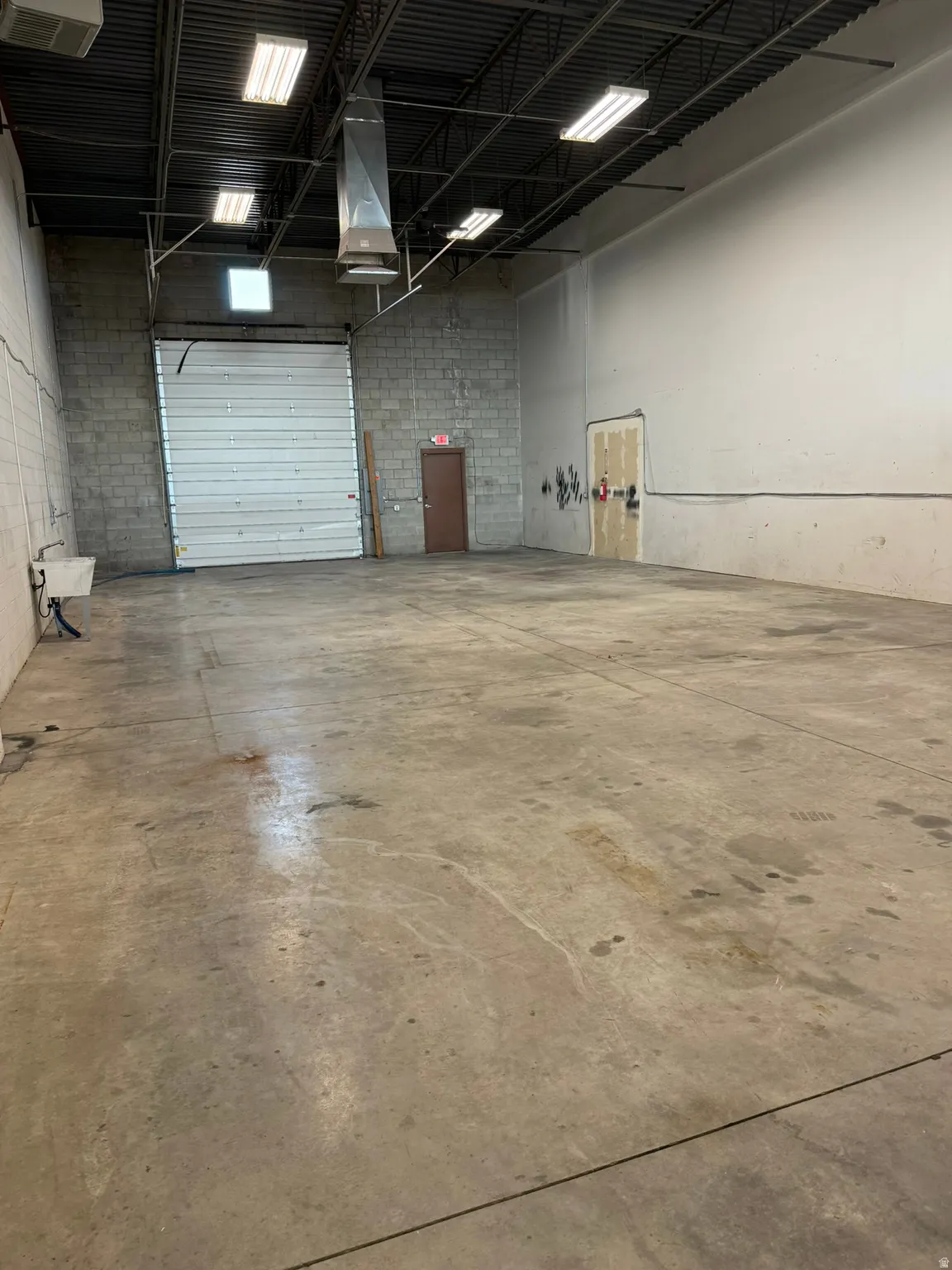 Open-concept warehouse space featuring a large roll-up door, exposed ceiling joists, concrete block wall, and rectangular fluorescent lighting fixtures