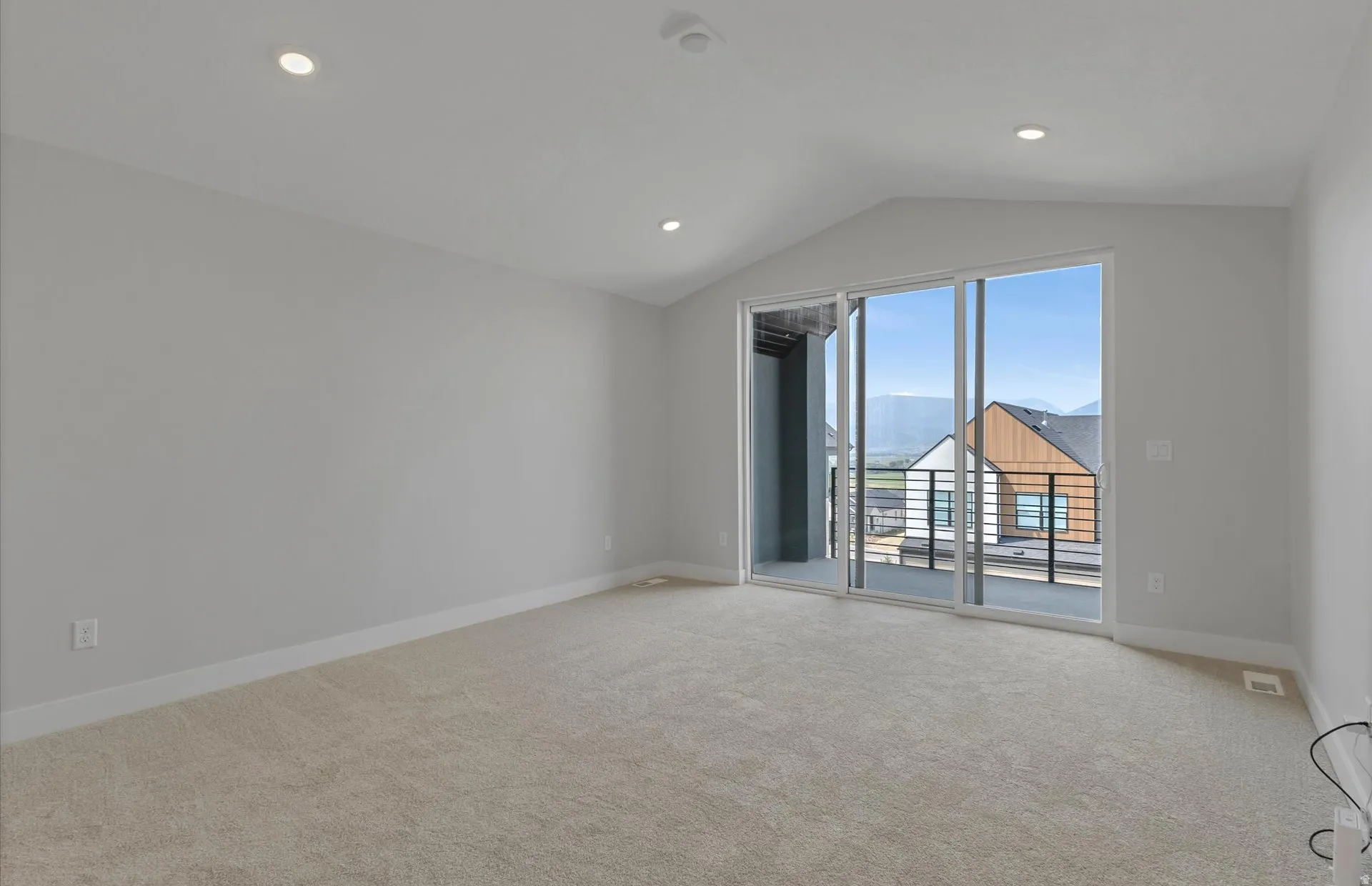 Vaulted ceiling with recessed lighting, light-toned carpeting, white baseboards, and a glass sliding door leading to a balcony with black metal railings