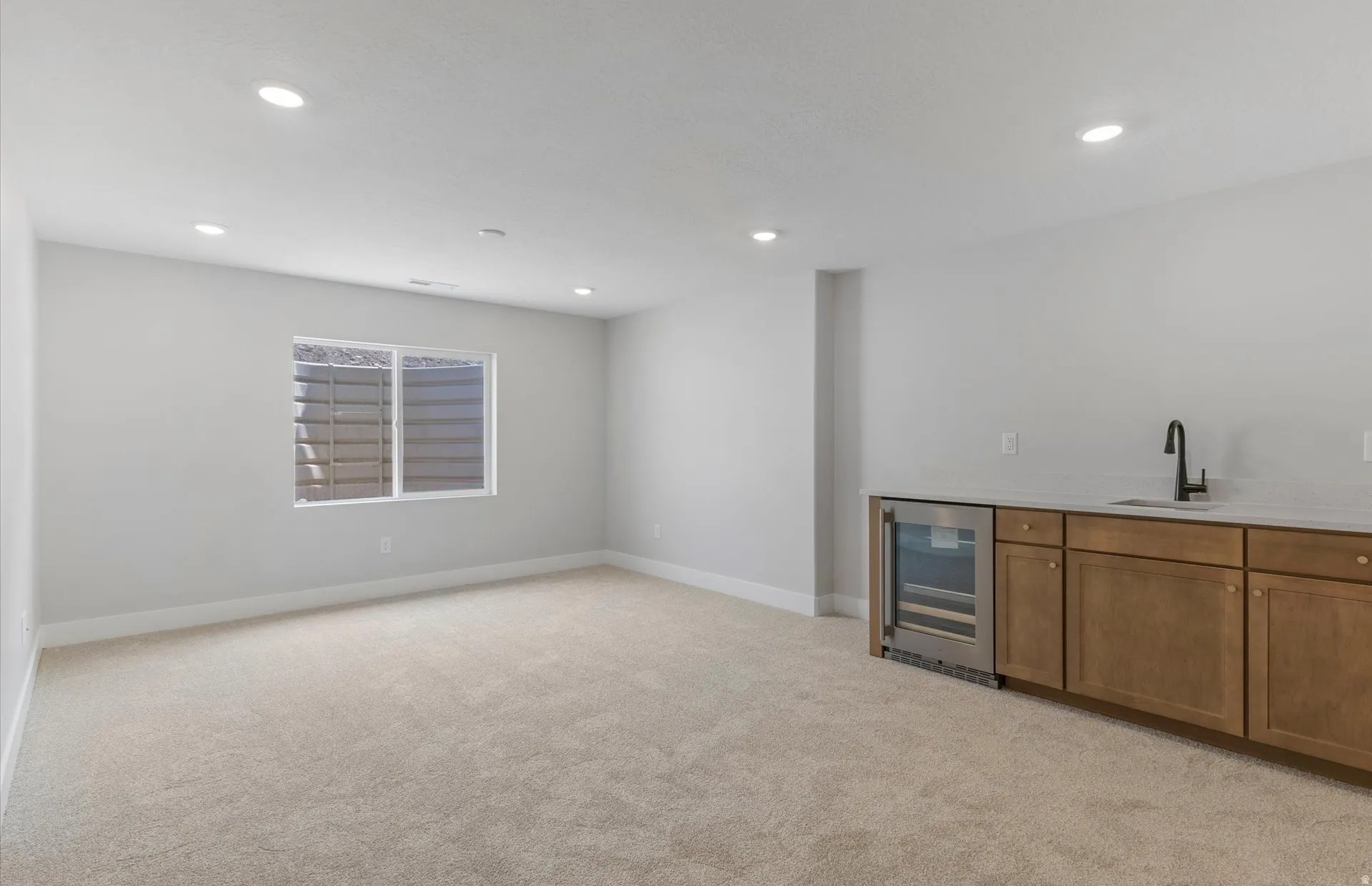 Recessed lighting illuminating a carpeted floor, window with exterior well, built-in cabinetry, under-counter beverage cooler, and a sink with a black fixture