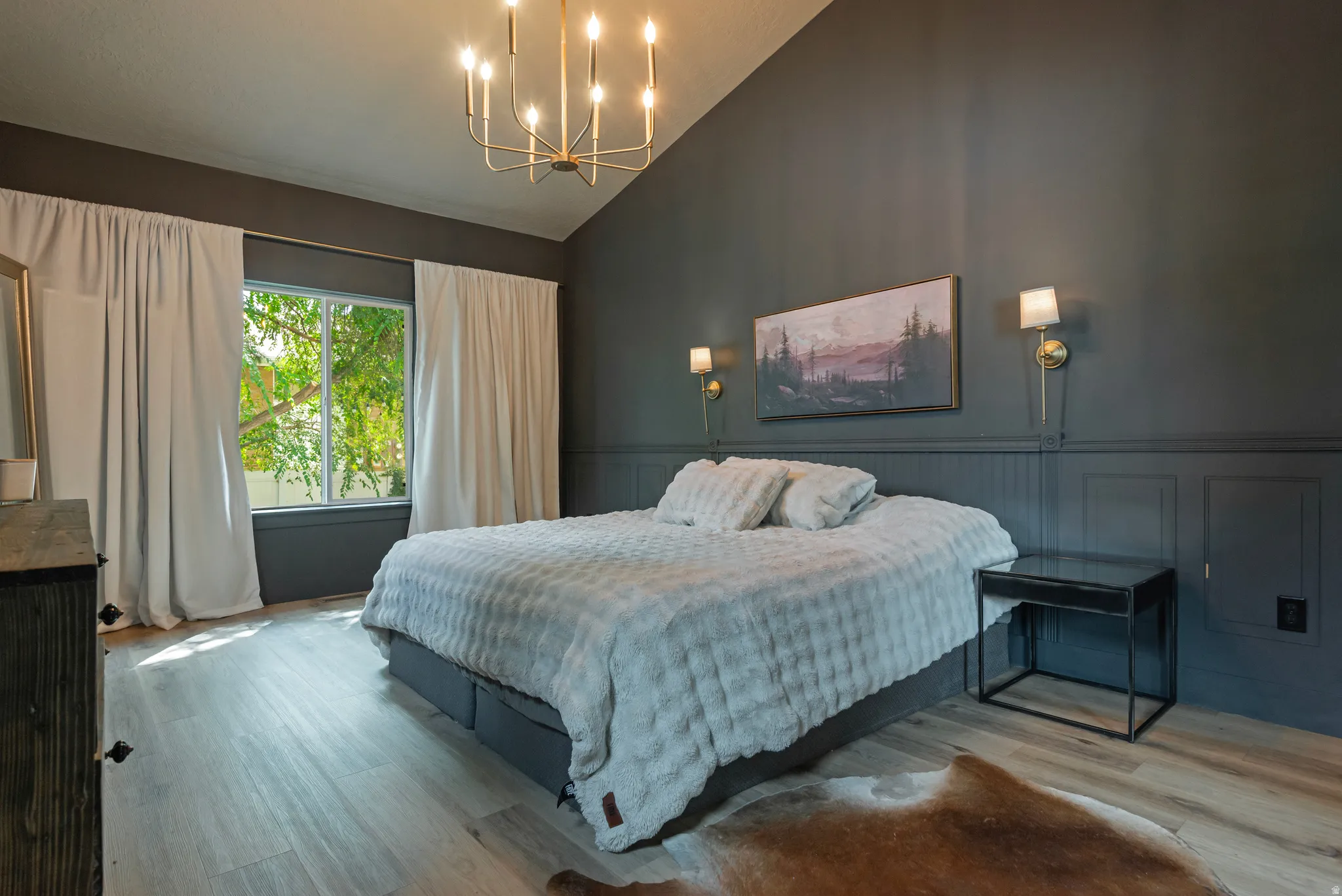 Sophisticated primary bedroom showcasing a custom wall paneling system with integrated lighting sconces, a modern chandelier fixture, and expansive windows with floor-to-ceiling drapery
