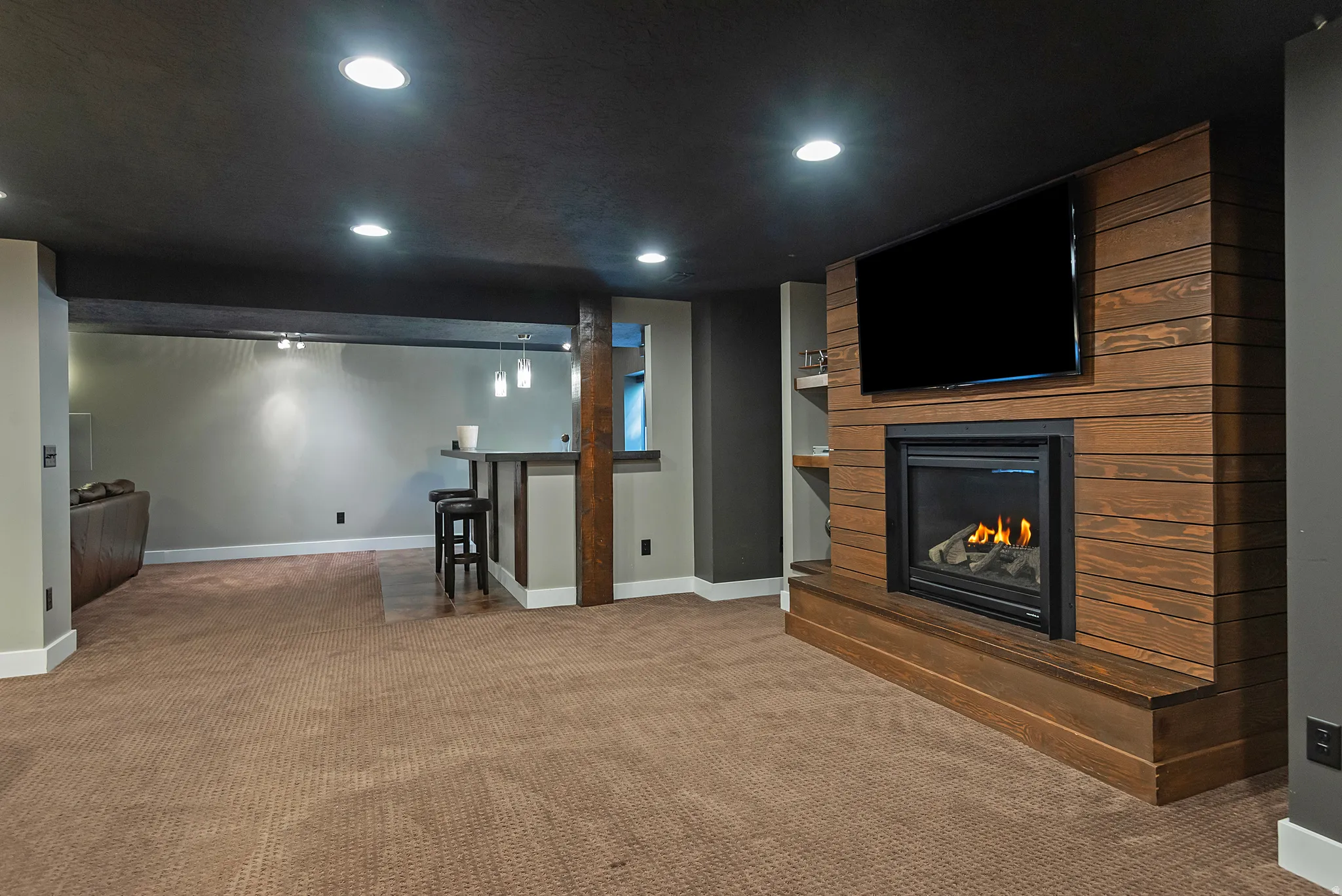 Spacious basement recreation area featuring recessed LED ceiling lighting, warm wood-finish wall paneling with an integrated gas fireplace and media nook, neutral carpet flooring, and multiple finished zones ideal for entertaining or personal use