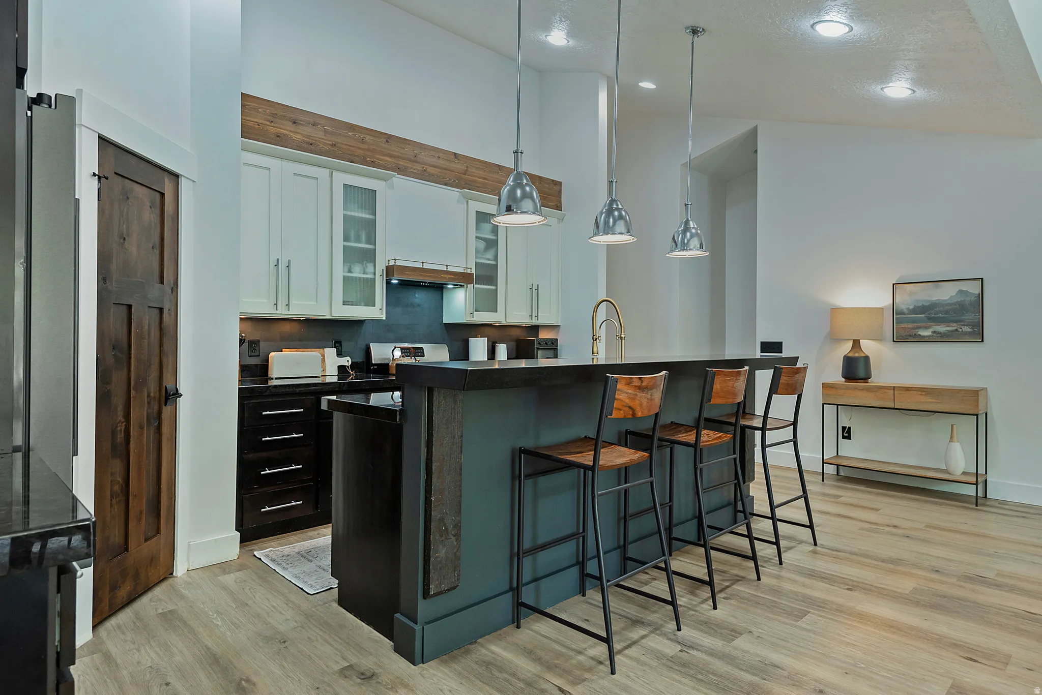 Kitchen featuring a central island with pendant lighting, white upper cabinetry with glass inserts, black lower cabinetry, wood-finish flooring, and a dark wood-paneled door